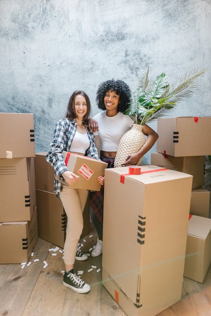 Two women stand inside a room during a home relocation process, surrounded by numerous packed cardboard boxes of various sizes, some sealed with red tape. One woman is holding a medium-sized box with