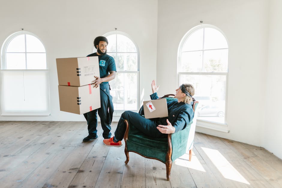 Two women stand inside a room during a home relocation process, surrounded by numerous packed cardboard boxes of various sizes, some sealed with red tape. One woman is holding a medium-sized box with